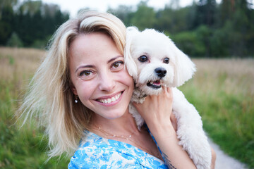 Portrait of smiling young woman kiss dog in Natural Park. Dog lover woman wearing blue dress with dog during good day. Concept of friendship and beautiful girl with a small dog