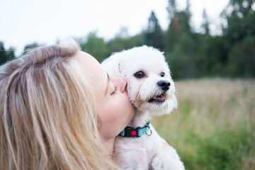 Portrait of smiling young woman kiss dog in Natural Park. Dog lover woman wearing blue dress with dog during good day. Concept of friendship and beautiful girl with a small dog