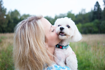 Portrait of smiling young woman kiss dog in Natural Park. Dog lover woman wearing blue dress with dog during good day. Concept of friendship and beautiful girl with a small dog