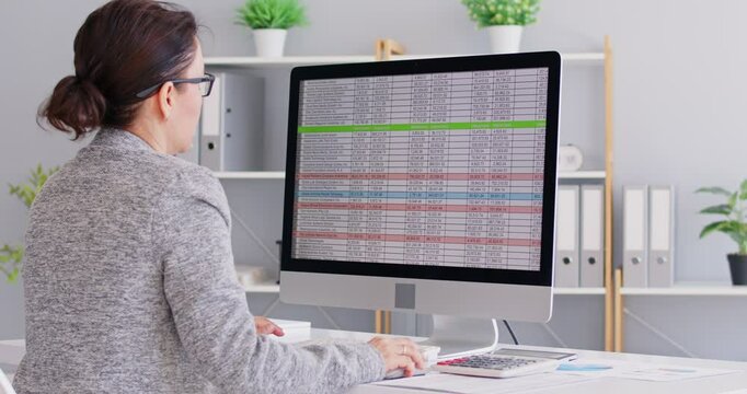 Back view portrait of a concentrated business woman accountant sitting at the desk and working on a pc computer with tables and charts at office and looking to the monitor screen. 4k video.