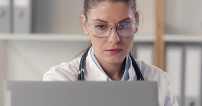 Young female online doctor, medical assistant working concentrated with laptop, wearing glasses. Woman performing medical consultant, healthcare administration, clinic management