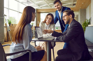 Young smiling business man showing work project to his colleagues on laptop computer while sitting at his workplace in office. Smiling people chatting on a meeting discussing company finance.