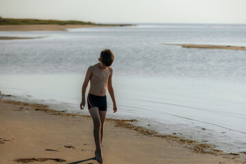 A young boy strolls barefoot along a quiet beach, looking down at the sand. The calm water stretches into the distance, creating a peaceful scene.