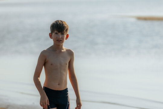 A young boy strolls barefoot along a quiet beach, looking down at the sand. The calm water stretches into the distance, creating a peaceful scene. - Powered by Adobe