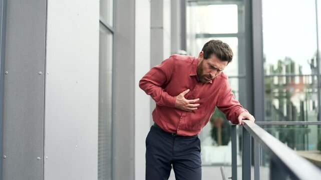 Man in red shirt suffered a heart attack while walking on street outside of office building. Upset bearded businessman suffers from tension in chest. He feels bad, experiences sharp pain in his chest