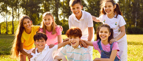 Happy children in a summer park. Outdoor group portrait of happy friends in a sunny green park. Seven kids sitting and standing on a green meadow, looking at the camera and smiling. Banner background