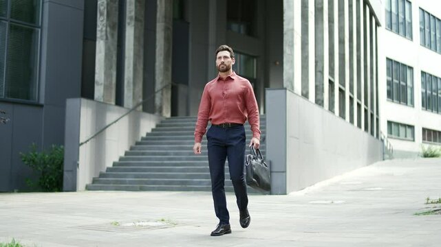 Businessman walking confidently outside modern office building with leather bag. Man smiling while enjoying sunny day in urban city setting. Business, entrepreneurship, and city life concepts