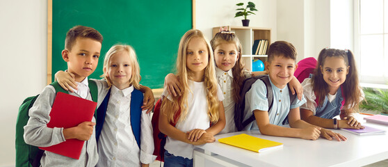 Portrait of a group of happy smiling primary school kids classmates standing in classroom and hugging after a lesson. Schoolchildren looking cheerful at camera. Education and back to school concept.