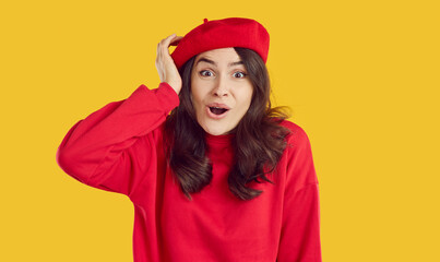 Photo of beautiful impressed young woman touching her head. Surprised brunette girl in red beret and sweatshirt staring at camera with open mouth over isolated on yellow background