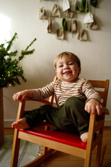 A smiling toddler sitting on a red chair, dressed in striped clothes, enjoying the moment. Christmas decorations and greenery in the background, creating a warm festive atmosphere. Isolated on a white