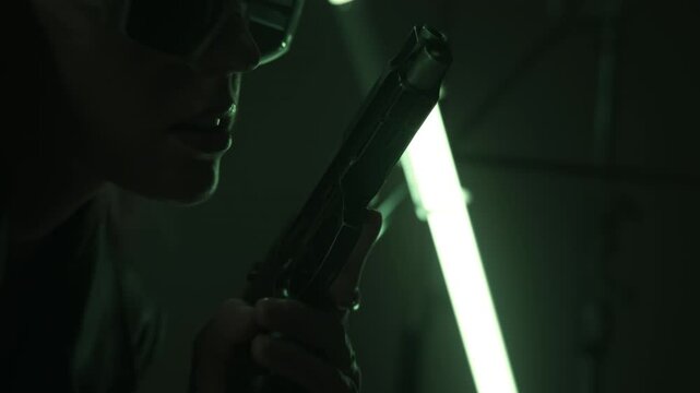 Beautiful futuristic lady in leather coat and sunglasses holding a gun and posing in a dark room lit by RGB tube lights