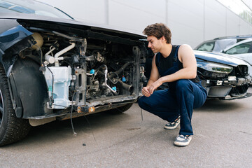 Pensive mechanic wearing blue overalls crouching next to damaged car, carefully examining for any necessary repairs, assessing plan for future work in automobile repair and renew service station.