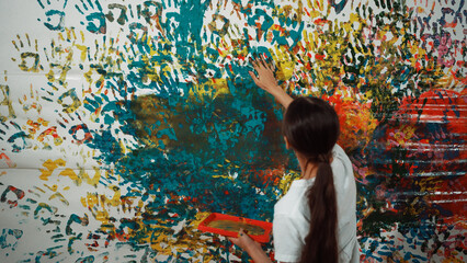 Naklejka premium Young happy girl holding the color tray and paint colorful stained wall to express idea and enhance imagination with hand print. Caucasian girl stamp hand to decorate room in art lesson. Edification.