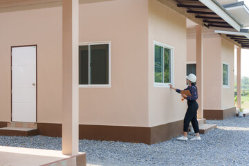 Female Asian building inspector wearing a hard hat standing outside of a house pointing at window...