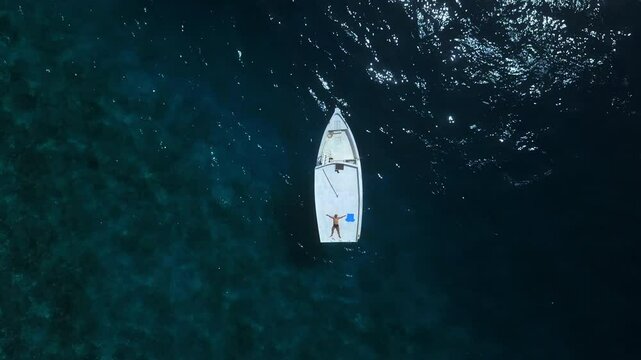 Aerial view on a man lies on the roof of a boat in ocean. Drone move down on a man on a wherry. Blue ocean with a shallop and a guy on top during  sunny day