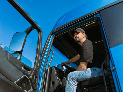 A young man is a professional driver in the cab of a blue truck. Handsome Caucasian guy, a transportation worker sits in a big truck car