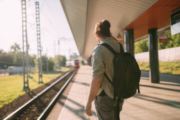 A man is waiting for a train on the platform. A young guy, a passenger with a backpack is standing on the platform waiting for the train