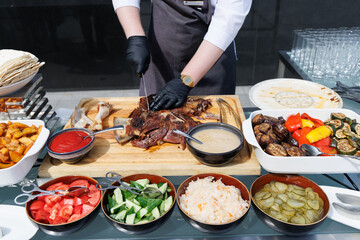 A chef is preparing food in front of a buffet table. The table is filled with various dishes, and the chef is using a spoon to serve food. Concept of busyness and anticipation