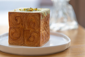 Close-up side view of freshly baked brown cube croissant with green pistachio cream on white plate on table in restaurant or cafe. Soft focus. Copy space. Puff pastry theme.
