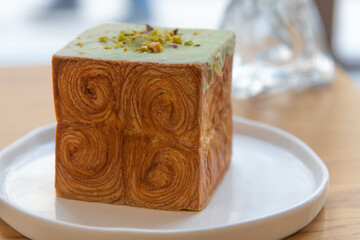 Close-up side view of freshly baked brown cube croissant with green pistachio cream on white plate on table in restaurant or cafe. Soft focus. Copy space. Puff pastry theme.
