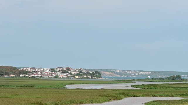 Coastal Wetlands near Igneada District. Scenic view of coastal wetlands with a winding river, grassy fields, and the distant town of Igneada under a clear sky.

