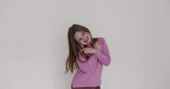 Cute little girl playfully touches face on white background. Child with pleasant smile expresses embarrassment posing for photo in studio
