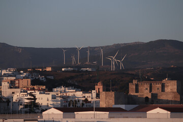 Fototapeta premium View of wind turbines on the mountains with curvy road between them, blue skies with white clouds at sunset with the city of Tarifa in the foreground