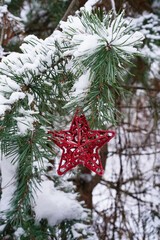 New Year, Christmas vertical template with red star for greeting card. Red shiny openwork star hanging on the branch of a fir tree covered with snow and ice in the forest, natural blurred background.