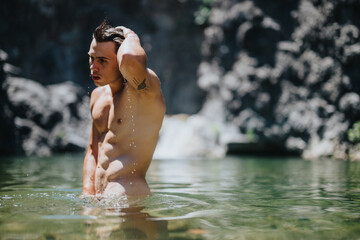 A young man stands waist-deep in a clear natural water pool, with rocky cliffs in the background, enjoying a refreshing dip.