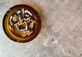 Brown glass ashtray full with cigarette butts close-up on grey stone background top view. Smoking kills.