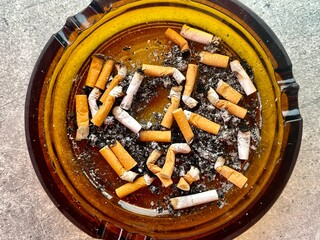 Brown glass ashtray full with cigarette butts close-up on grey stone background top view. Smoking kills.