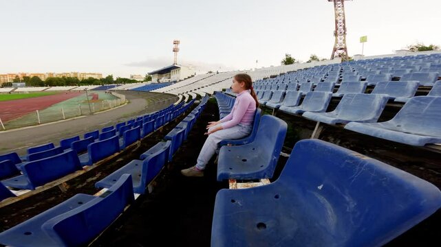Young girl sits alone in an empty stadium, waiting and watching. Changing positions, she reflects on her surroundings, embodying a sense of solitude and anticipation