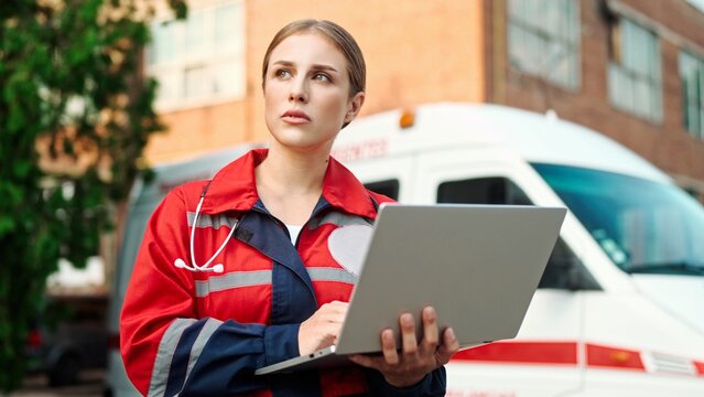 Beautiful Caucasian young medical worker woman in red uniform typing on laptop surfing internet standing on street near hospital. Close up of female paramedic using computer outdoor near ambulance