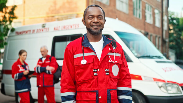 Joyful smiling African American male doctor in red medical uniform looking at camera standing outdoor near ambulance vehicle. Paramedics with tablet device on background, first aid, healthcare concept