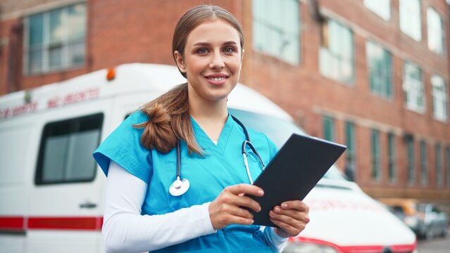 Caucasian pretty young professional medical worker using tablet standing in street near ambulance car. Close up portrait. Beautiful female nurse in blue uniform tapping on device and smiling - Powered by Adobe