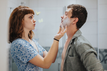 A young couple in the bathroom getting ready for a night out, with the woman helping the man shave. They appear playful and focused on grooming.