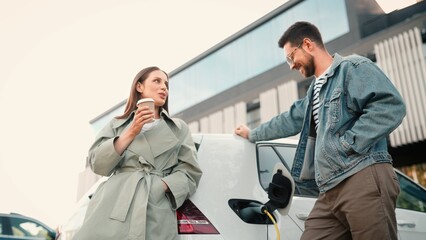 Happy young couple stand outside in city and talking waiting till vehicle charge. Happy woman drinking coffee while man putting cable in electric car at charger station. EV charging. Zerro emission