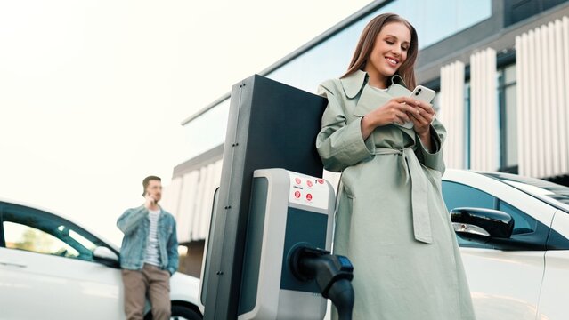 Joyful smiling young pretty woman charging her electric car standing outside at charging point and texting on smartphone. People charge electrocars at charger station outdoors. Low angle view
