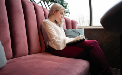 Dreamy young woman writing down something in her own diary resting in cozy cafeteria