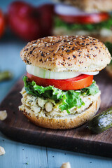 A vegan vegetarian sandwich made with chickpea spread, lettuce, slices of tomatoes and onions with pickles on the side over everything bagel seasoned buns. Selective focus with blurred background.