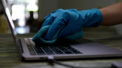 Close-up hands of unrecognizable woman wearing blue glove carefully cleaning laptop keyboard with green microfiber cloth, promoting thorough disinfection and hygiene in dark office environment.