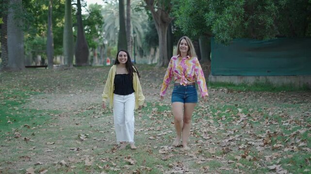 two young caucasian girls one brunette and one blonde walking barefoot on the grass towards the camera making thumbs up gestures and so-so gestures