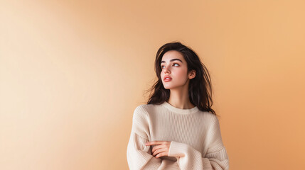Obraz premium Beautiful girl in a warm jumper against a beige wall points her finger sideways. Dark-haired girl in cosy jumper, copy space on the side