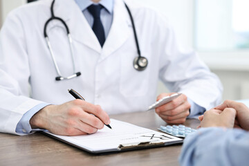 Doctor with pills writing prescription for patient at wooden table in clinic, closeup