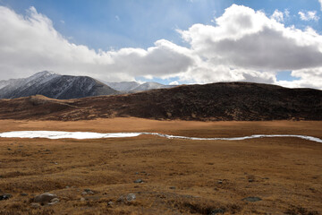 The frozen bed of a shallow winding river crosses the flat steppe along the mountain range on a sunny autumn day.
