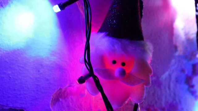 Una toma de cerca de una decoraci&oacute;n de navidad y fin de a&ntilde;o con una cara de santa claus  rodeado de varias luces con bombillos de varios colores en movimiento en la oscuridad colocadas en una pared 