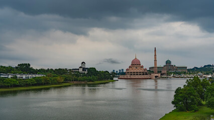 A calm lake in the city park. Green vegetation on the banks. A beautiful pink mosque is visible in the distance. Domes and minaret against a cloudy sky. Malaysia. Putrajaya. Putra Mosque 