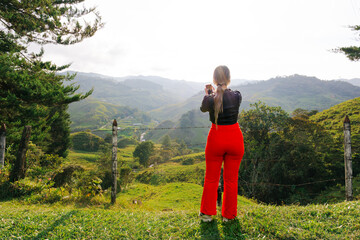 Fototapeta premium Traveling woman in red pants taking a photo in nature surrounded by trees. Road.