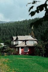 A charming two-story red house surrounded by lush greenery in a mountainous region on a cloudy day