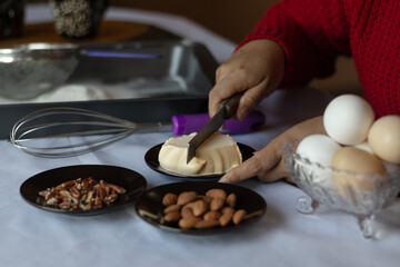 Close-up of a woman's hands making a dessert, cutting butter with a knife, next to ingredients such as eggs, almonds and walnuts on a table, preparing it for baking.